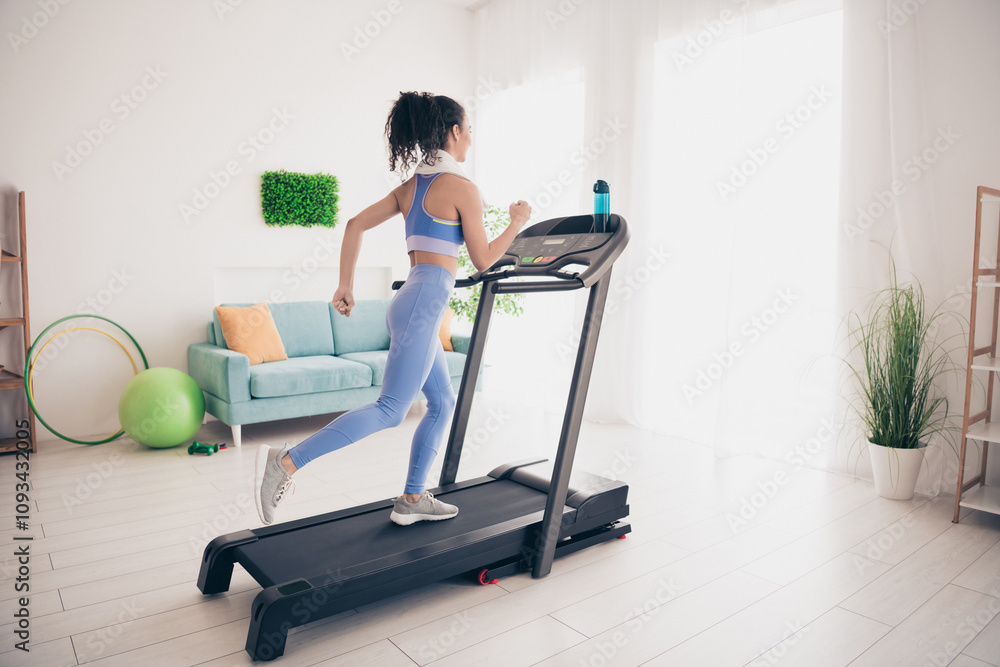 Fototapeta premium Young woman exercising on treadmill in modern apartment with sunlight and vibrant decor, promoting healthy lifestyle and indoor fitness.