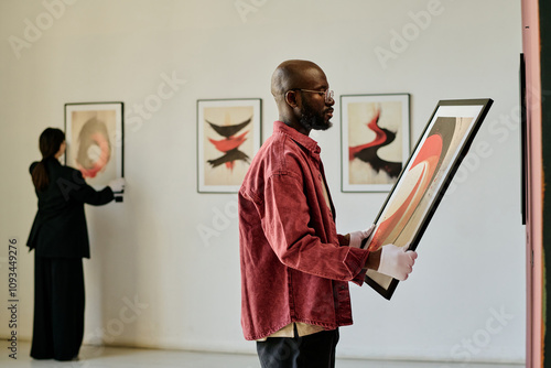 African American man holding abstract painting, curating art gallery exhibit. Background reveals another person examining framed artwork on white walls
