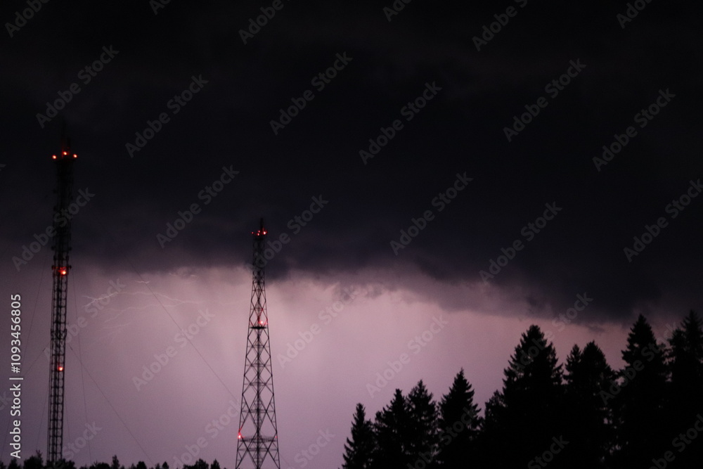 Naklejka premium Lightning during a night thunderstorm near a forest and two television towers.