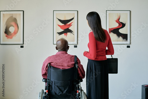 Two people observing abstract artworks displayed on wall in modern art gallery. One person in wheelchair appreciating paintings up close photo