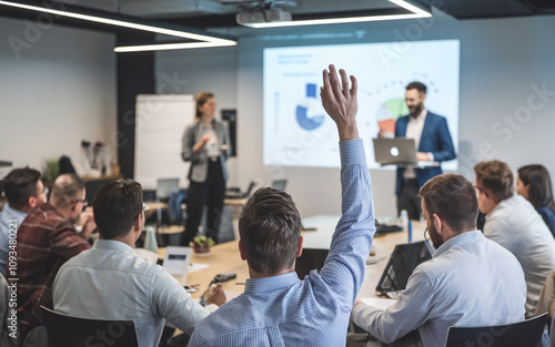 Group of participants in an interactive business seminar, with one individual raising their hand while seated and another person presenting in the background with a laptop.

