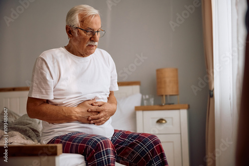 Senior man holding his abdomen in pain while sitting on  bed. photo