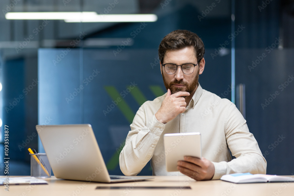 A serious young man businessman sits in the office at the table with a laptop, holds his hand by his beard and looks thoughtfully at the tablet in his hand A serious young man businessman sits in the office at the table with a laptop, holds his hand by his beard and looks thoughtfully at the tablet in his hand