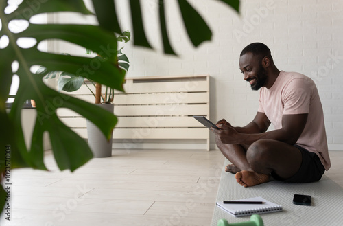 Young African-American man sitting on a yoga mat at home after exercising, smiling while using a tablet for a wellness app in a bright, plant-filled space