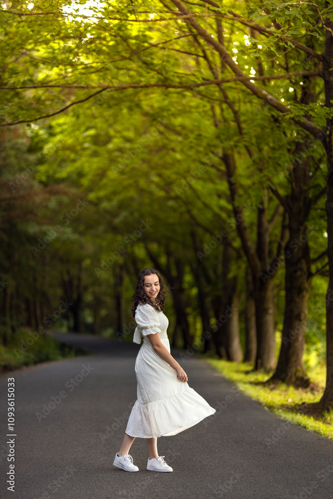 Joyful smiling dancing girl in white dress with sneakers on a dirt road among trees. Laughing woman in a summer park in a tree alley Joyful smiling dancing girl in white dress with sneakers on a dirt road among trees. Laughing woman in a summer park in a tree alley