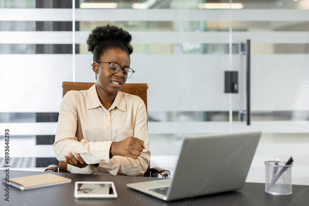 Obraz premium Businesswoman feels arm pain while working at a laptop in modern office. Woman in glasses, formal attire supports elbow joint, indicating discomfort. Contemporary workspace fosters productivity