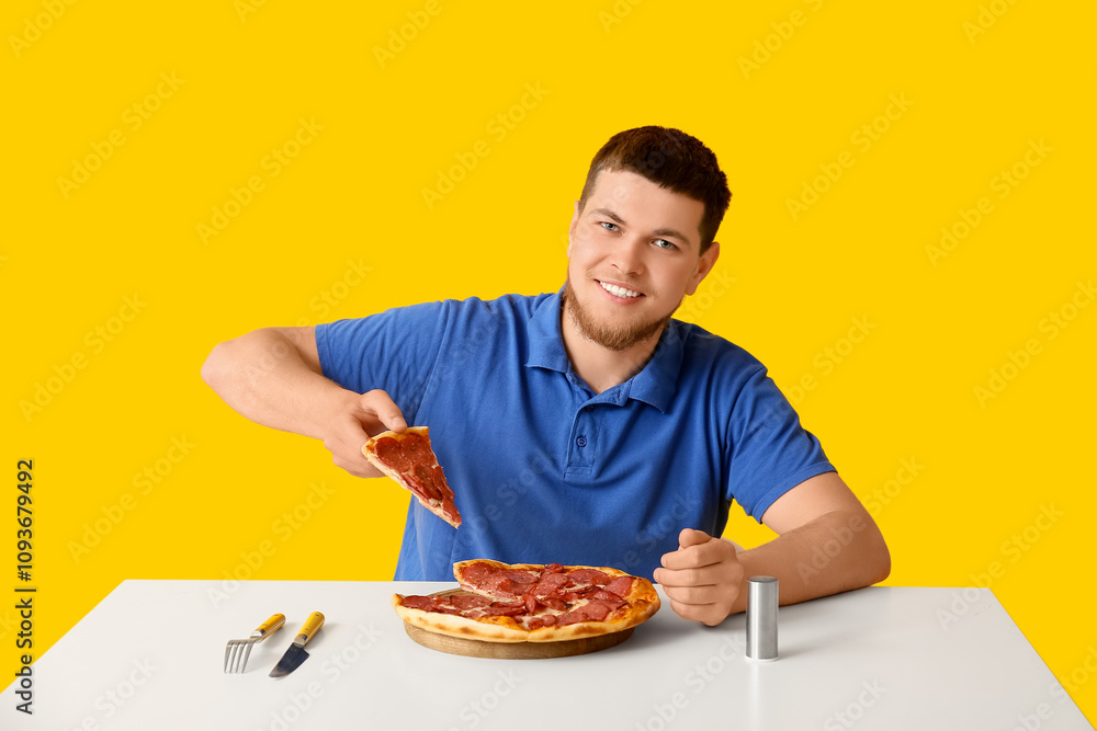 Young man with tasty pepperoni pizza at table on yellow background Young man with tasty pepperoni pizza at table on yellow background