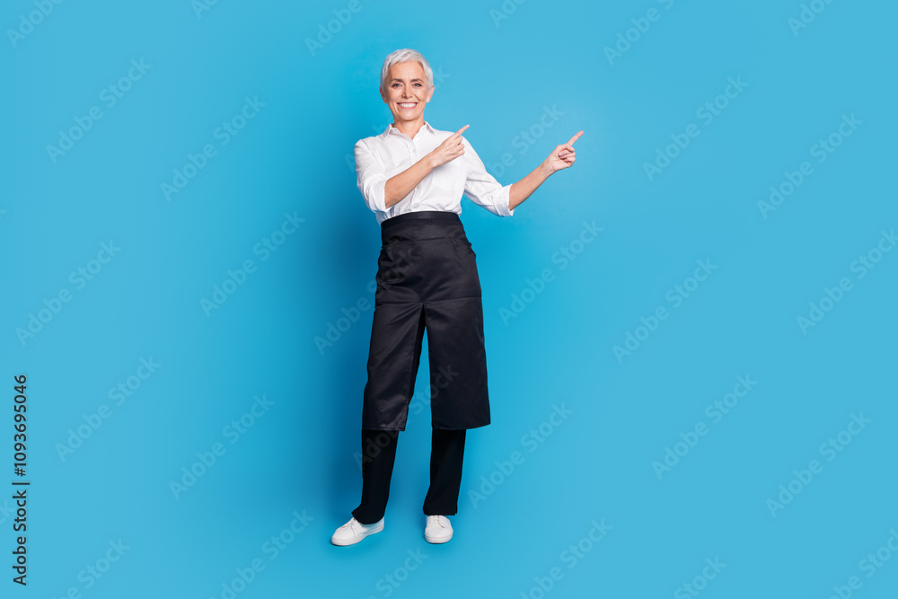 Full body photo of attractive senior woman small business cafe owner point empty space dressed waitress uniform isolated on blue background Full body photo of attractive senior woman small business cafe owner point empty space dressed waitress uniform isolated on blue background