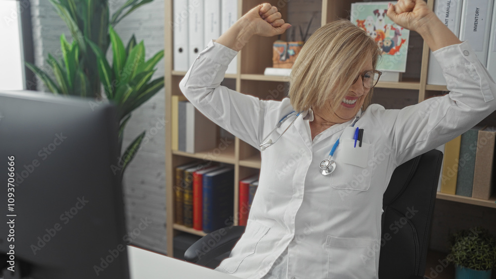 Obraz premium A smiling middle-aged blonde woman in a clinic office, wearing a white medical coat and stethoscope, raises her arms in excitement.