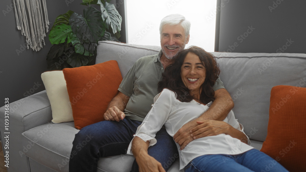Middle-aged couple smiling together on a cozy sofa in a stylish living room with plants and soft cushions, showcasing a loving relationship in a comfortable home setting. Middle-aged couple smiling together on a cozy sofa in a stylish living room with plants and soft cushions, showcasing a loving relationship in a comfortable home setting.