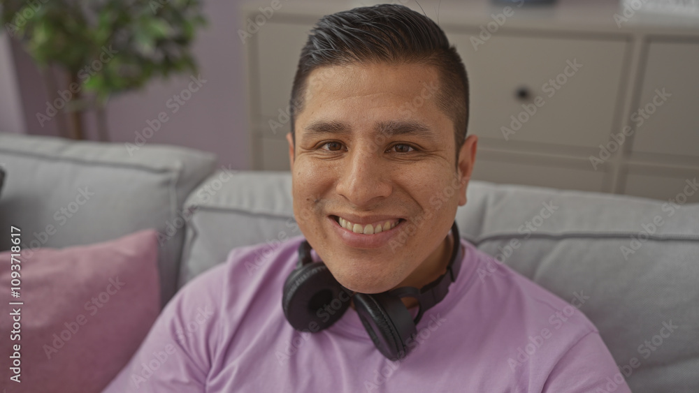 A smiling young hispanic man with headphones around his neck, posing in a cozy living room setting. A smiling young hispanic man with headphones around his neck, posing in a cozy living room setting.