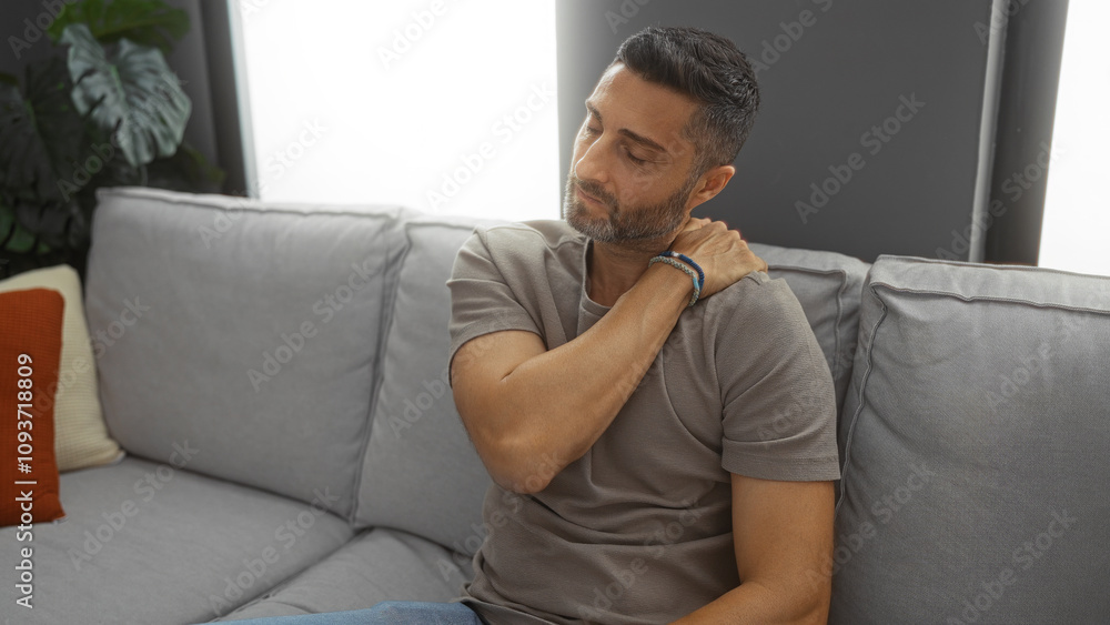 Man sitting on a couch in a modern living room, appearing exhausted while massaging his neck, sunlight streaming through a window illuminating the cozy interior. Man sitting on a couch in a modern living room, appearing exhausted while massaging his neck, sunlight streaming through a window illuminating the cozy interior.