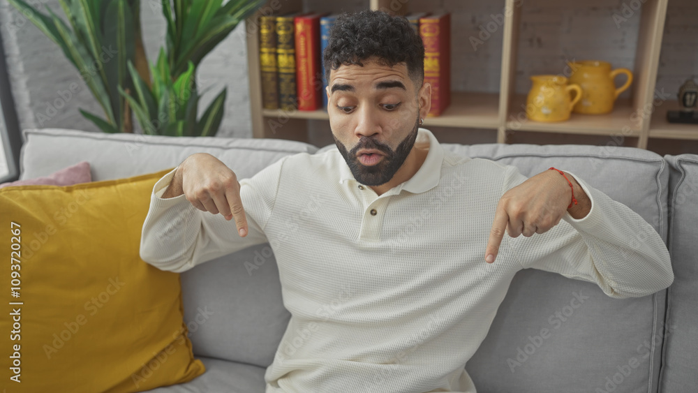 Fototapeta premium Surprised hispanic man with beard sitting on couch indoors pointing downwards with a playful expression