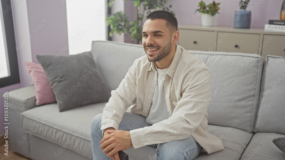 Smiling young hispanic man sitting comfortably on a gray sofa at home in a well-lit modern living room Smiling young hispanic man sitting comfortably on a gray sofa at home in a well-lit modern living room