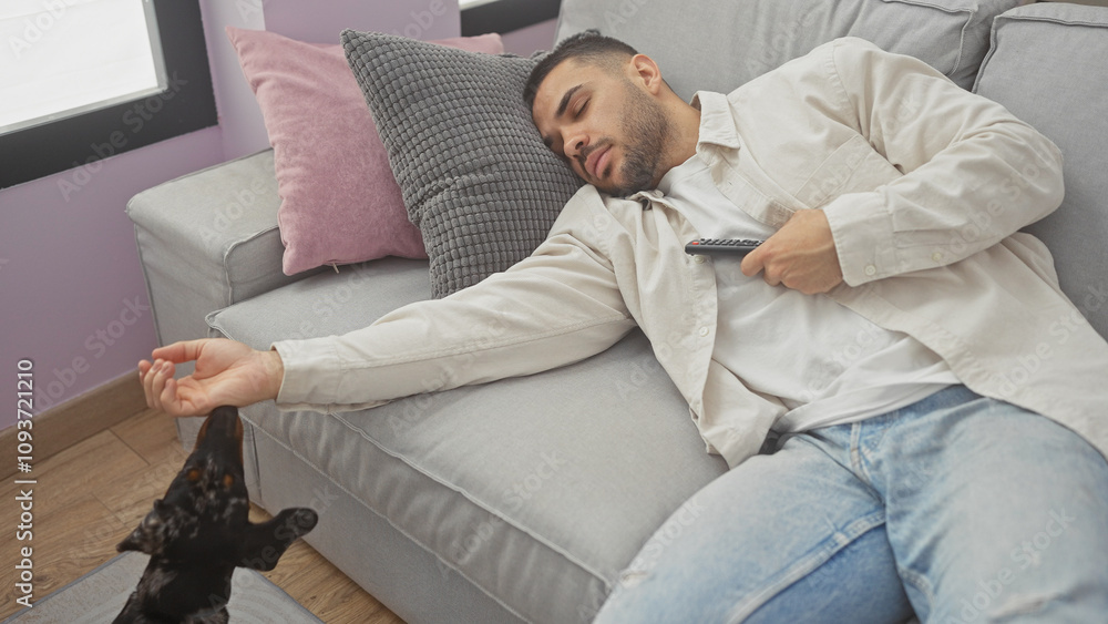 A young hispanic man sleeps on a sofa holding a tv remote with a teckel dog at his side in a cozy living room. A young hispanic man sleeps on a sofa holding a tv remote with a teckel dog at his side in a cozy living room.