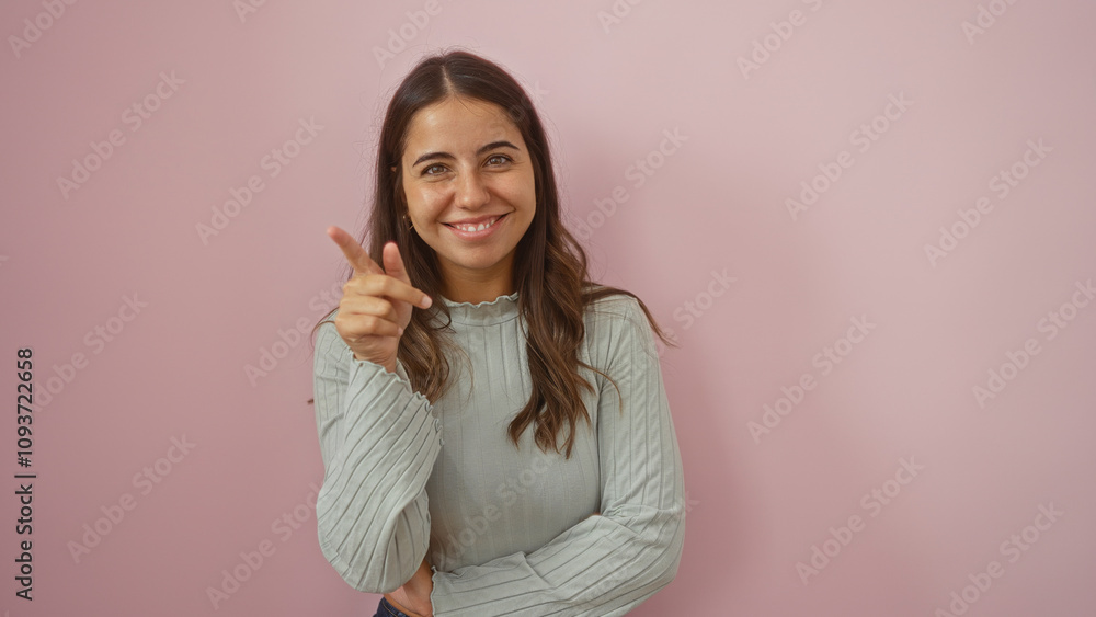 Woman pointing smiling with playful expression over pink background wall showcasing confident young adult with brunette hair in casual clothing approachable and beautiful. Woman pointing smiling with playful expression over pink background wall showcasing confident young adult with brunette hair in casual clothing approachable and beautiful.