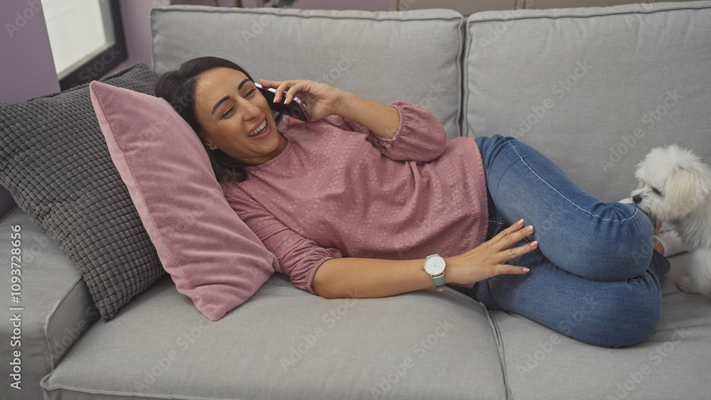 Obraz premium A smiling woman lying on a couch talking on the phone with a maltese dog beside her in a modern living room.
