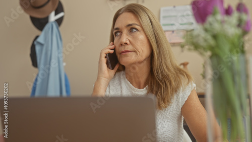 A middle-aged woman converses on a phone in an indoor home setting, with flowers and a laptop visible.