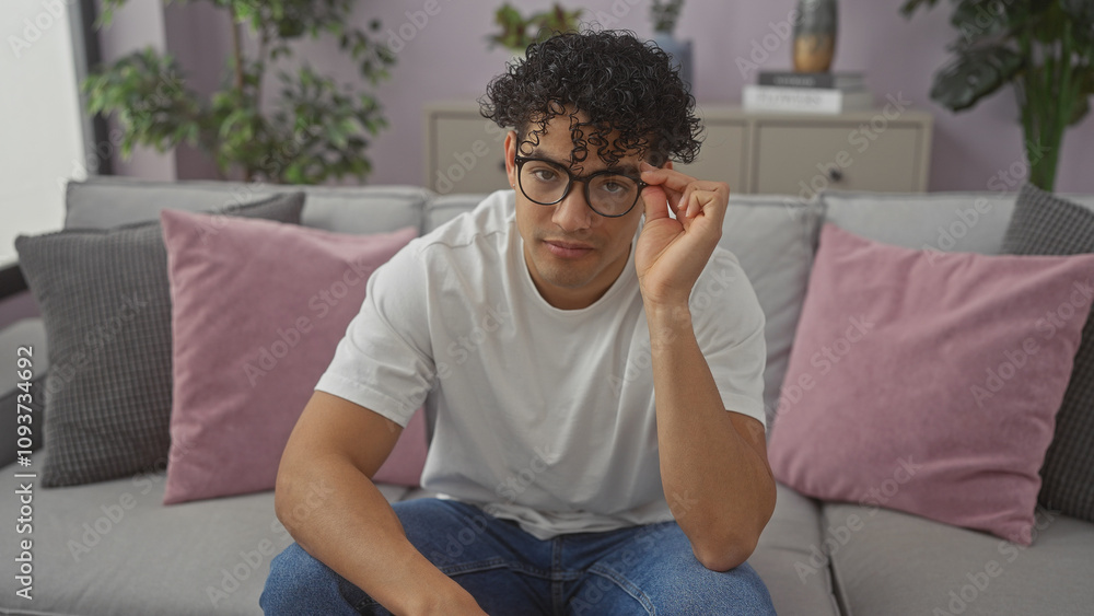 A young hispanic man in casual clothing sitting thoughtfully on a couch indoors, adjusting his glasses. A young hispanic man in casual clothing sitting thoughtfully on a couch indoors, adjusting his glasses.