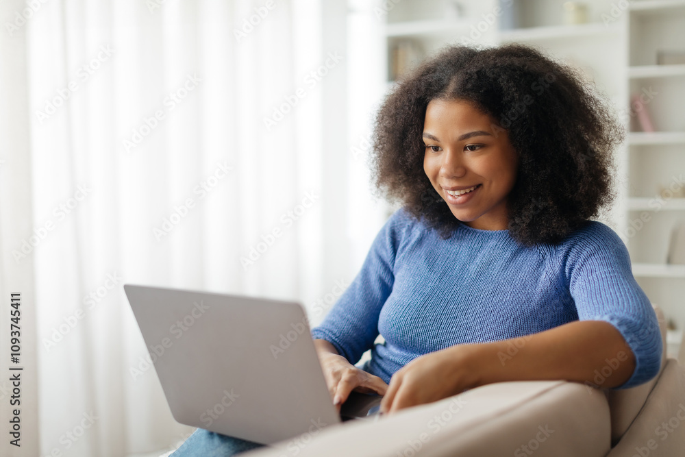 Cheerful young black woman using laptop while sitting on sofa at home, smiling african american female freelancer browsing or working online on computer, typing on keyboard, copy space. High quality Cheerful young black woman using laptop while sitting on sofa at home, smiling african american female freelancer browsing or working online on computer, typing on keyboard, copy space. High quality
