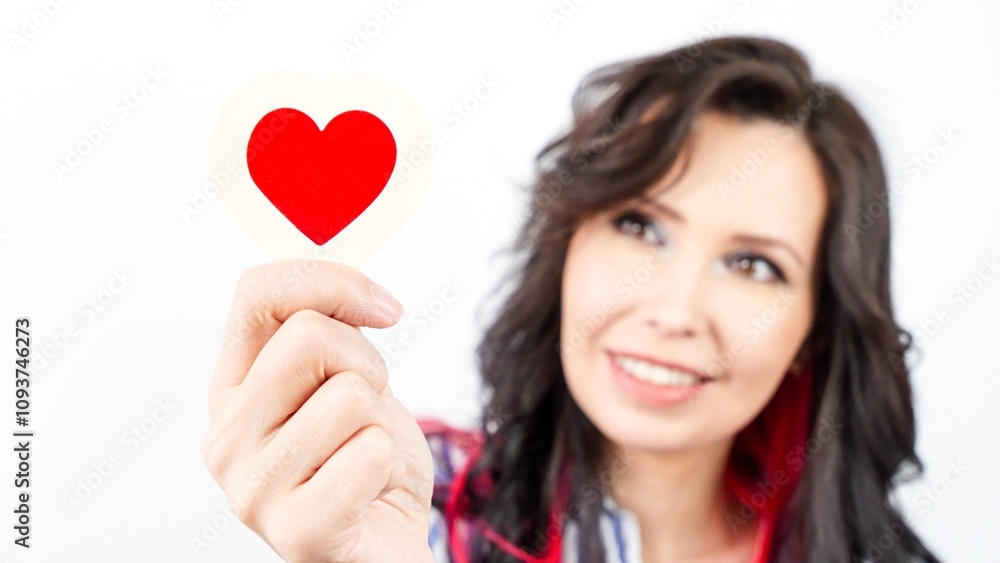Cheerful young woman with long dark hair joyfully holding a red heart-shaped cutout in her hand. Cheerful young woman with long dark hair joyfully holding a red heart-shaped cutout in her hand.