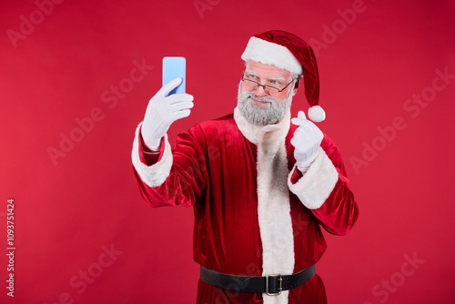Tableau sur toile Elderly man in Santa Claus outfit taking selfie with blue smartphone on red background