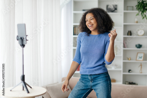 Happy Young Black Woman Dancing At Camera While Recording Video At Home, Positive African American Female Creating Content For Social Networks, Using Smartphone On Tripod In Living Room, Copy Space