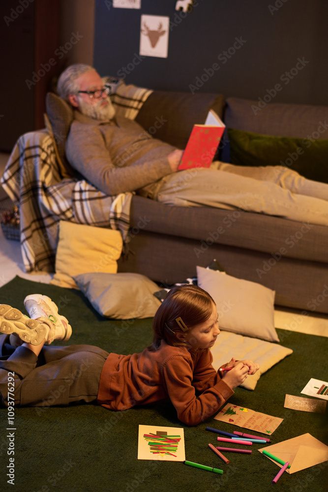 Fototapeta premium Senior man relaxing on couch reading book with child lying on floor drawing. Cozy home environment with pillows scattered around room creating warm atmosphere