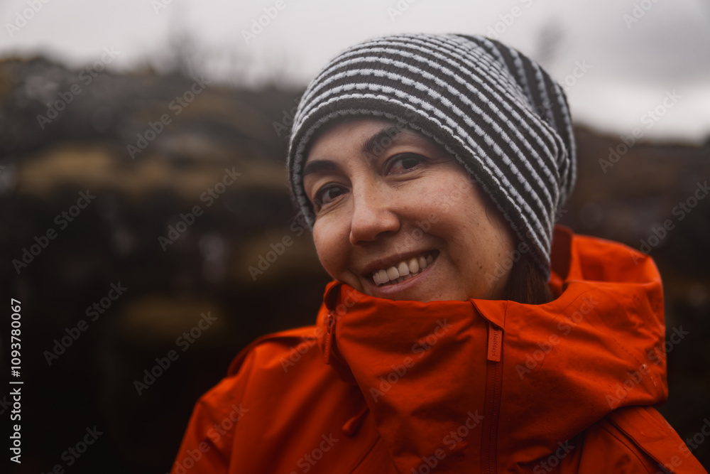 Portrait of a smiling hiker wearing an orange jacket and beanie against a blurred mountainous backdrop Portrait of a smiling hiker wearing an orange jacket and beanie against a blurred mountainous backdrop