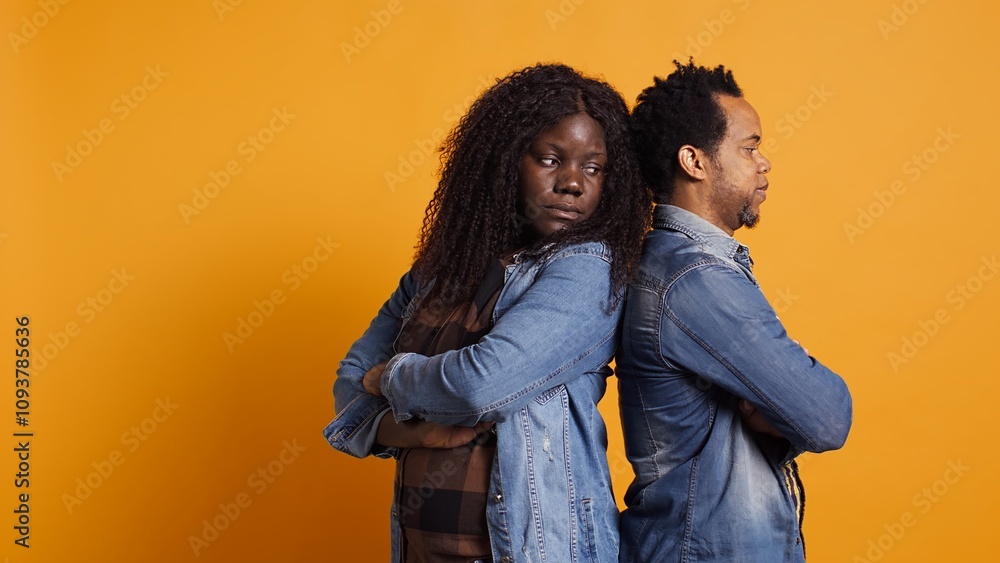 Fototapeta premium Distant sad husband and wife regret fighting each other in studio, standing back to back after a misunderstanding. African american couple with marriage problems, quarreling. Camera B.