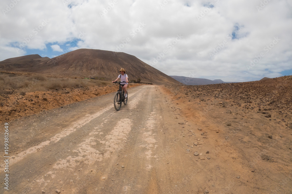 a middle aged woman in a cowboy hat riding a bicycle through a desertic landscape, horizontal, La Graciosa, Lanzarote a middle aged woman in a cowboy hat riding a bicycle through a desertic landscape, horizontal, La Graciosa, Lanzarote