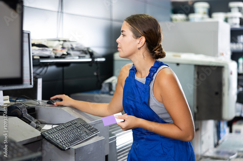 Young woman employee of paint and construction materials warehouse stands near computer and looks at screen, checks input data for collecting an online order