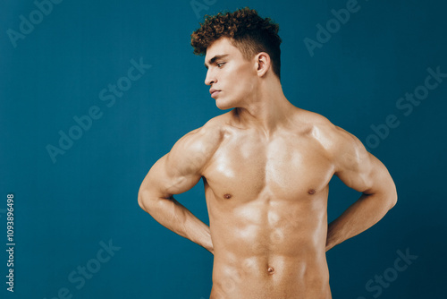 Confident young man with curly hair, standing shirtless against a blue background. Fitness and health concept, showcasing body positivity and self-acceptance.