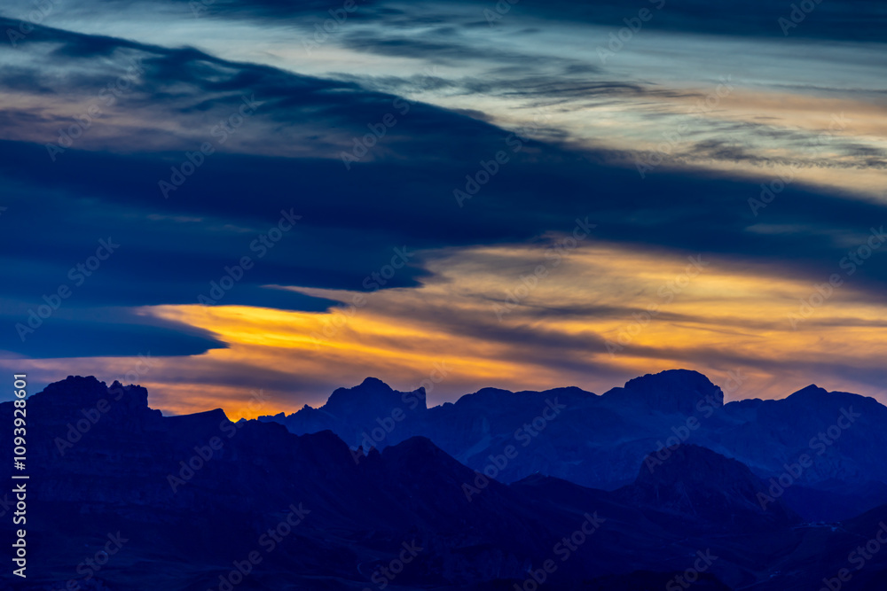 Fototapeta premium Sunset in the mountains. Red and orange burning sky horizon behind the alpine mountain range. Rocky mountain peaks of the Dolomites at sunset and dawn at sunrise light sunrays going through the clouds