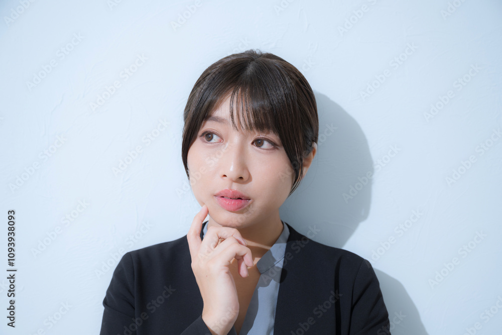 Business scene of troubled young woman wearing suit and thinking with hand on chin, light blue background, frontal view of troubled job change or job hunting. Business scene of troubled young woman wearing suit and thinking with hand on chin, light blue background, frontal view of troubled job change or job hunting.