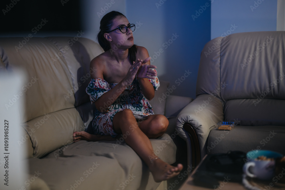 Obraz premium A woman relaxes on a comfortable sofa, immersed in a television show. She appears captivated and content, enjoying a calm evening at home with a cozy indoor ambiance.