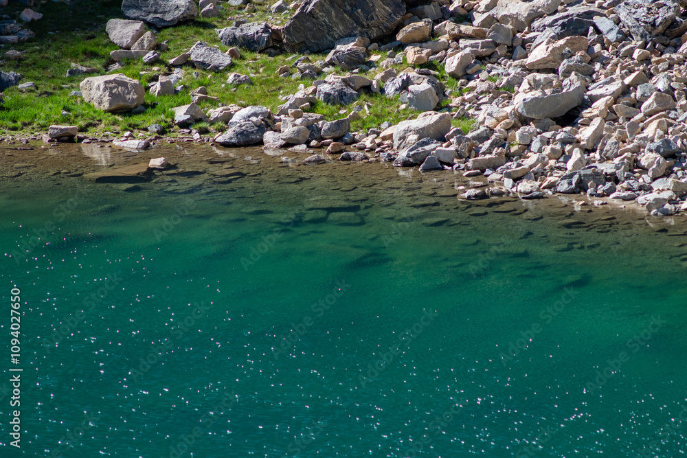 Clear turquoise water gently laps against the rocky shore of a serene lake surrounded by greenery on a sunny day Clear turquoise water gently laps against the rocky shore of a serene lake surrounded by greenery on a sunny day