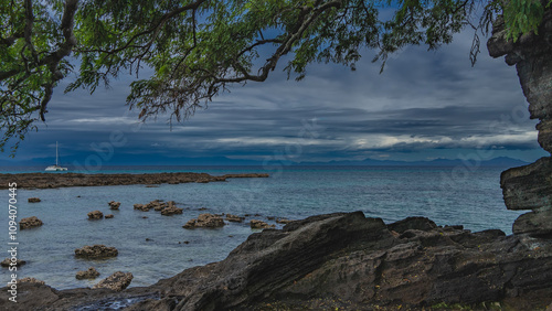 The branches of the tree bent over the rocky shoreline. Clams attached to boulders exposed at low tide. In the turquoise ocean a white yacht. Mountains against a cloudy sky. Madagascar. Nosy Tanikely photo