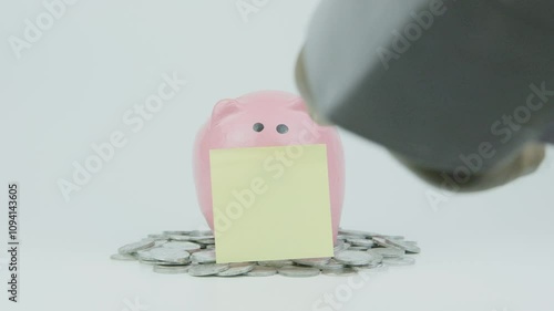 Hand Putting Yellow Note Paper to Eye of Pink Piggy Bank and Coin on White Background