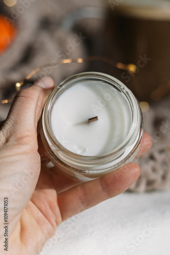 A woman's hand holds a candle with a wooden wick against a background of lights and a knitted plaid. Vertical photo