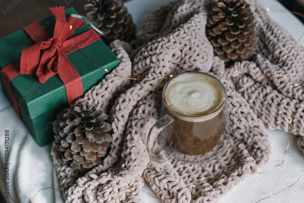 Winter still life. A glass cup of coffee with milk, big cones, lights, a green gift box with a red bow. Modern christmas rustic eco friendly decor, scandinavian winter hygge. Atmospheric christmas