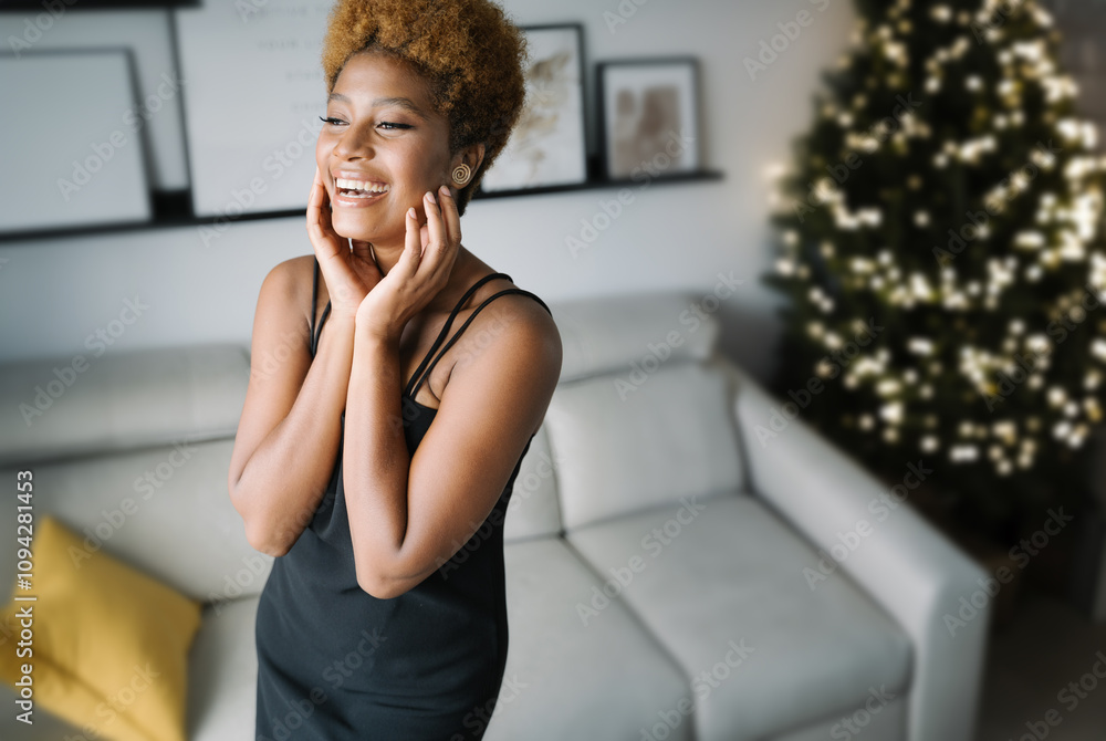 Fototapeta premium Happy laughing black woman in elegant little dress in cozy light interior decorated Christmas tree. Winter holidays magic moment. Closing eyes and touching her face with her hands. Wow emotions