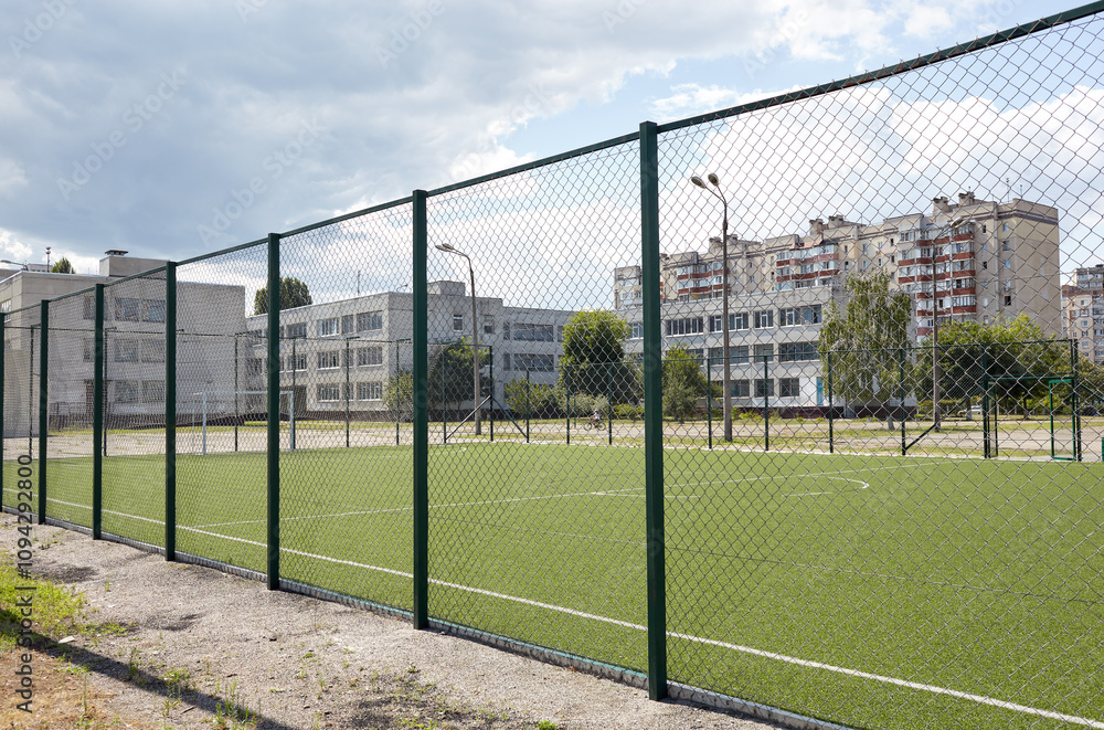 Fototapeta premium Lawn field for playing football behind the green fence mesh at school territory. Close-up of soccer field with green grass