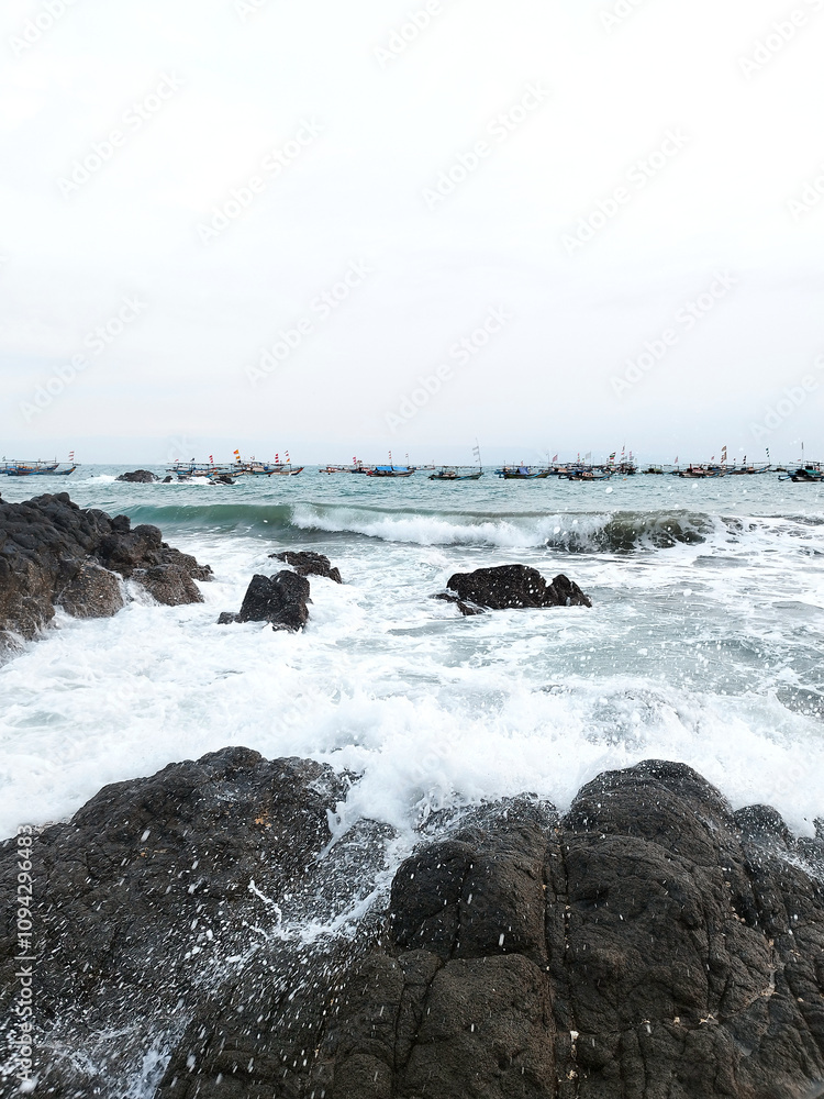 View of beautiful beach, with Sea bedrock. Fishermen boats and sky horizon at the background, near sunset time. Dark sediment rock  sea banks, wave, water splashing, and black sand.