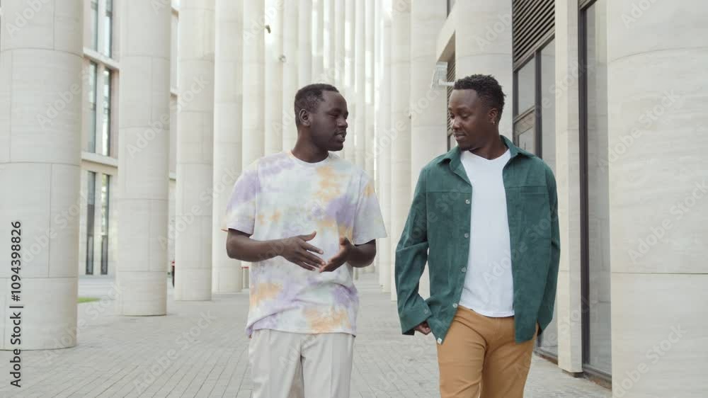 Two African American male students walk along campus buildings, talking. Co workers on break chill outdoors chatting. Cheerful black skinned friends enjoy communication smiling Two African American male students walk along campus buildings, talking. Co workers on break chill outdoors chatting. Cheerful black skinned friends enjoy communication smiling