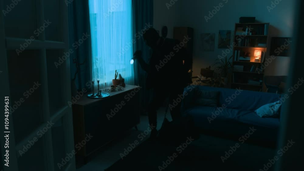Unknown criminal dressed in black clothes with bag stealing jewellery, candle holders and tray from dark room while his partner checking bookcase Unknown criminal dressed in black clothes with bag stealing jewellery, candle holders and tray from dark room while his partner checking bookcase