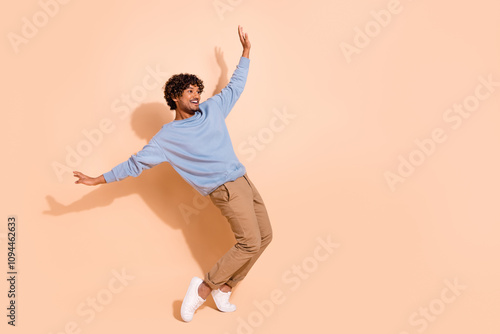 Full body photo of carefree guy wear blue shirt trousers stand on tiptoes look at sale empty space isolated on beige color background