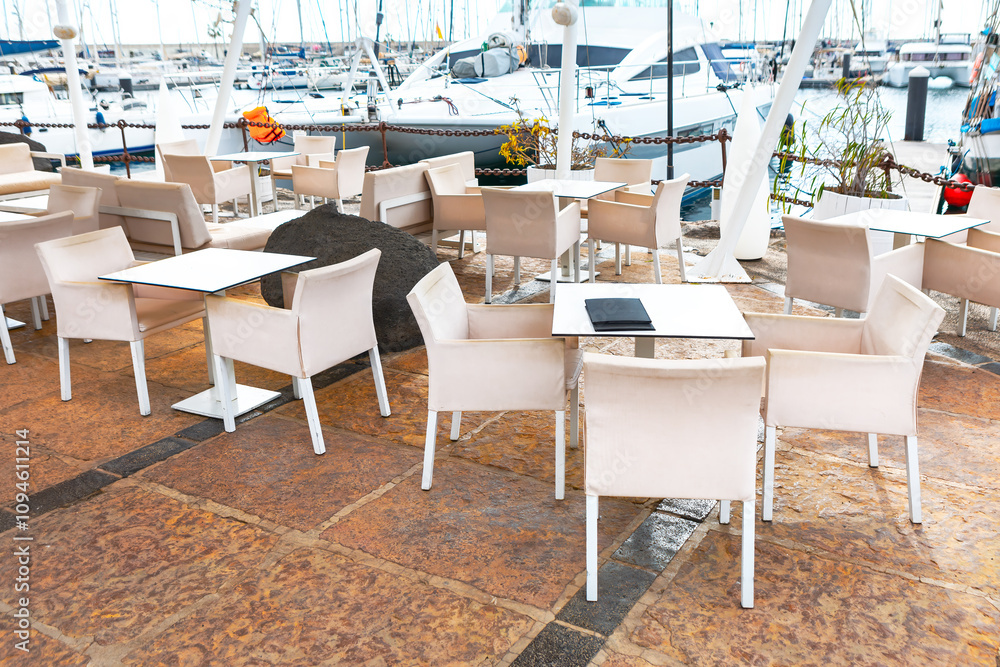 Outdoor seating area at a marina, with tables and chairs arranged on a tiled floor. In the background, there are boats docked at the marina, adding a scenic and nautical atmosphere to the setting