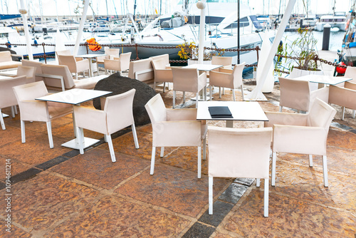 Obraz na płótnie Outdoor seating area at a marina, with tables and chairs arranged on a tiled floor. In the background, there are boats docked at the marina, adding a scenic and nautical atmosphere to the setting w salonie