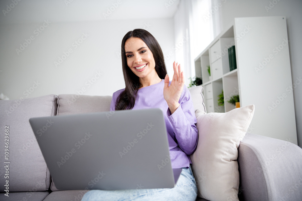 Charming young woman sitting on the sofa with a laptop, waving happily during an online call in a stylish living room setting Charming young woman sitting on the sofa with a laptop, waving happily during an online call in a stylish living room setting
